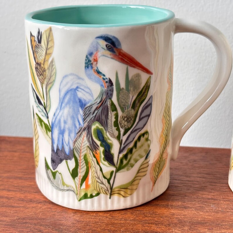 Puede incluir: Taza de cer&aacute;mica blanca con una ilustraci&oacute;n de una garza azul. La taza tiene un interior turquesa y un asa curva. La garza est&aacute; representada en tonos azules, con un pico rojo, rodeada de follaje colorido.