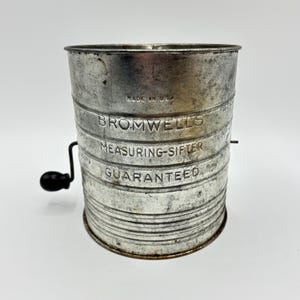 Puede incluir: Un vaso medidor vintage de metal plateado con una manivela manual. El vaso tiene las palabras "Bromwell's Measuring-Sifter Guaranteed" impresas en el lateral.