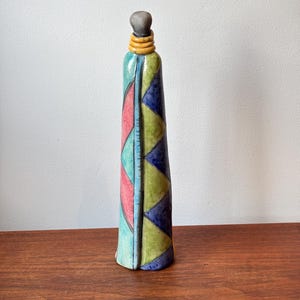 May include: A tall, slender ceramic figurine with a rounded grey head and yellow neck rings. The body features a geometric pattern of triangles in shades of pink, light blue, lime green, and dark blue, separated by thin black lines.