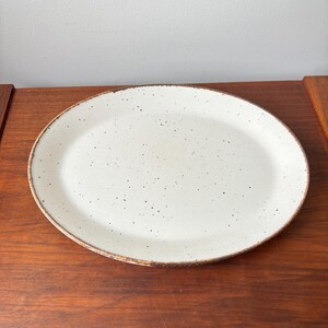 Puede incluir: Plato ovalado de cerámica con una superficie blanca moteada y un borde marrón. El plato está sobre una superficie de madera. Este plato es ideal para servir aperitivos, entrantes o postres.