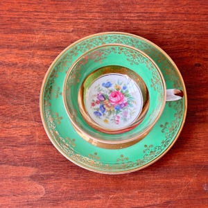 Könnte beinhalten: Ein Vintage-Teeservice in Grün und Gold. Das Set besteht aus einer Teetasse, einer Untertasse und einer kleinen Schale, die alle ein zentrales Blumenmuster aufweisen. Die Teetasse und die Untertasse sind mit Gold verziert. Das Set steht auf einer Holzoberfläche.