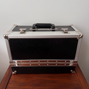 Puede incluir: Maletín de maquillaje negro con ribetes y herrajes plateados. El maletín tiene un asa negra y la marca "MAC" es visible en la parte superior. El maletín está sobre una superficie de madera.