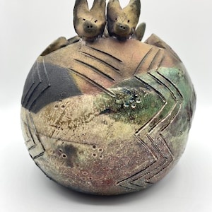 Może przedstawiać: Ceramiczny wazon w kształcie kuli z fakturowanym, wielokolorowym szkliwem. Na górze wazonu znajdują się dwie małe ceramiczne figurki zwierząt.