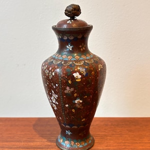 May include: An antique vase with a lid, featuring a detailed floral design. The vase is a rich reddish-brown, with patterns in white, blue, and gold. The lid has a textured knob. The vase is displayed on a wooden surface.