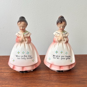 May include: Two ceramic figurines of figures with brown hair, wearing pink dresses with white aprons featuring blue and yellow floral accents. The left figurine has the text "Give us this day our daily bread" and the right has "We give you thanks for all your gifts".