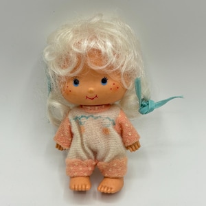 May include: A small, white doll with blonde hair wearing a pink and white sleeper. The doll has blue eyes and a painted smile.