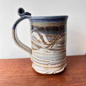 May include: Handmade ceramic mug with a distinctive handle. The mug has a mix of blue, brown, and white hues, with a textured finish. The handle has a decorative swirl at the top. A rustic, handcrafted piece for enjoying hot drinks.