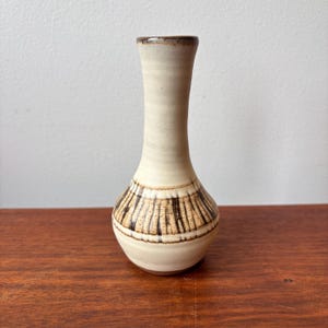 May include: A small, cream-coloured ceramic vase with a narrow neck and a rounded base. The vase features a band of vertical brown lines around the middle. The rim is a darker brown. The vase is displayed on a wooden surface.