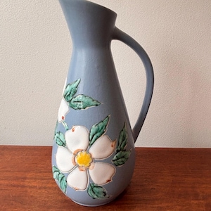 Puede incluir: Jarra de cerámica azul con asa y diseño floral. La jarra presenta una flor blanca con centro amarillo y hojas verdes. La jarra mide aproximadamente 25 cm de altura.