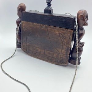 Pode incluir: Uma caixa de madeira com um desenho entalhado de uma casa e duas figuras nos lados. A caixa tem uma alça e uma figura de madeira entalhada no topo.