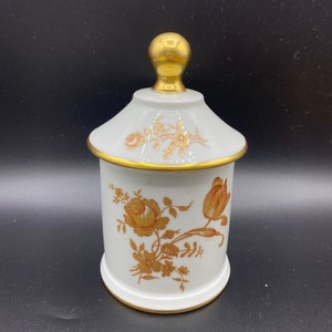 May include: White porcelain jar with a gold lid and floral design. The jar has a gold trim around the top and bottom. The flowers are in shades of brown and gold.