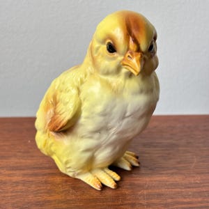 May include: A ceramic chick figurine in yellow with brown detailing on its head and wings. The chick has black eyes and a yellow beak. The figurine is displayed on a wooden surface.