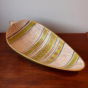 May include: A leaf-shaped serving dish in a mid-century modern style. The dish has a textured beige background with curved stripes in green, black, and gold. The rim is gold, and it sits on a dark wooden surface. The dish is approximately 30 cm long.
