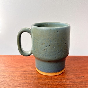 May include: A handmade ceramic mug in shades of blue and green with a textured surface. The mug has a cylindrical shape with a handle and a contrasting orange-yellow base. The mug is approximately 4 inches tall.
