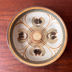 May include: A round ceramic plate with a cream-colored center and a brown rim. The plate features a symmetrical design of brown and tan floral-like shapes and lines radiating from the center. The plate is set on a dark wooden surface.