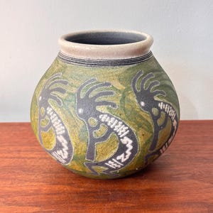 May include: A round ceramic vase with a green background and Kokopelli figures in black and white. The vase has a beige rim with black stripes and sits on a wooden surface.