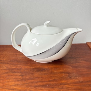 May include: White ceramic teapot with a curved handle, lid, and spout. The teapot has a black and grey striped design on the side. The teapot is on a wooden surface.