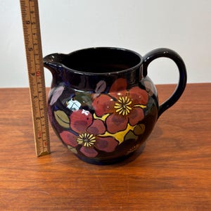 Op de afbeelding: Een donkerblauwe keramische kan met een handvat en tuit. De kan is versierd met rode en gele bloemmotieven. Een houten liniaal is naast de kan geplaatst en toont de afmetingen in centimeters.