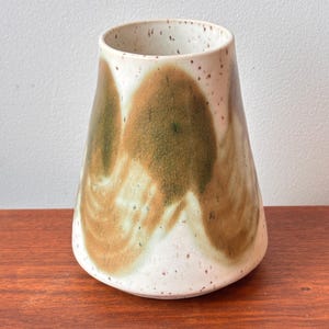 Może przedstawiać: Ręcznie wykonany ceramiczny wazon z nakrapianą białą podstawą i rozszerzanym kształtem. Wazon posiada abstrakcyjne brązowe i zielone pociągnięcia pędzla, tworzące unikalny, artystyczny design. Odpowiedni do eksponowania kwiatów lub jako element dekoracyjny.