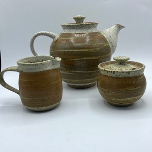May include: A brown and white ceramic teapot, creamer, and sugar bowl set with a speckled glaze and brown stripes.