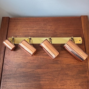 Pode incluir: Um conjunto de quatro copos medidores cilíndricos de cobre de tamanhos variados, cada um com uma pequena alça, suspensos em um suporte de parede horizontal de latão com quatro ganchos e um orifício de montagem na extremidade direita.