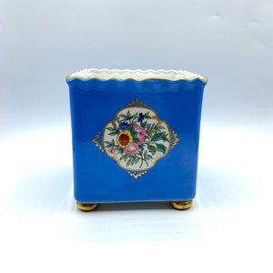 Op de afbeelding: Een kleine, vierkante, blauwe keramische plantenpot met een gegolfde rand en gouden accenten. De pot heeft een bloemenmotief met roze, gele en witte bloemen in een gouden frame.