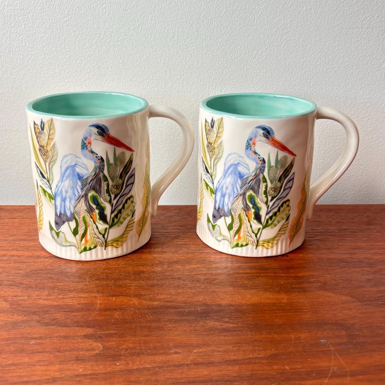 Puede incluir: Dos tazas de cer&aacute;mica con exterior crema e interior verde agua claro. Cada taza presenta una ilustraci&oacute;n detallada de una garza azul con pico rojo, rodeada de follaje verde y amarillo. Las tazas tienen un asa curva.