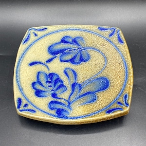 Op de afbeelding: Een vierkante keramische tegel met een blauw en wit bloemenmotief. De tegel heeft een getextureerd oppervlak en een licht verhoogde rand.