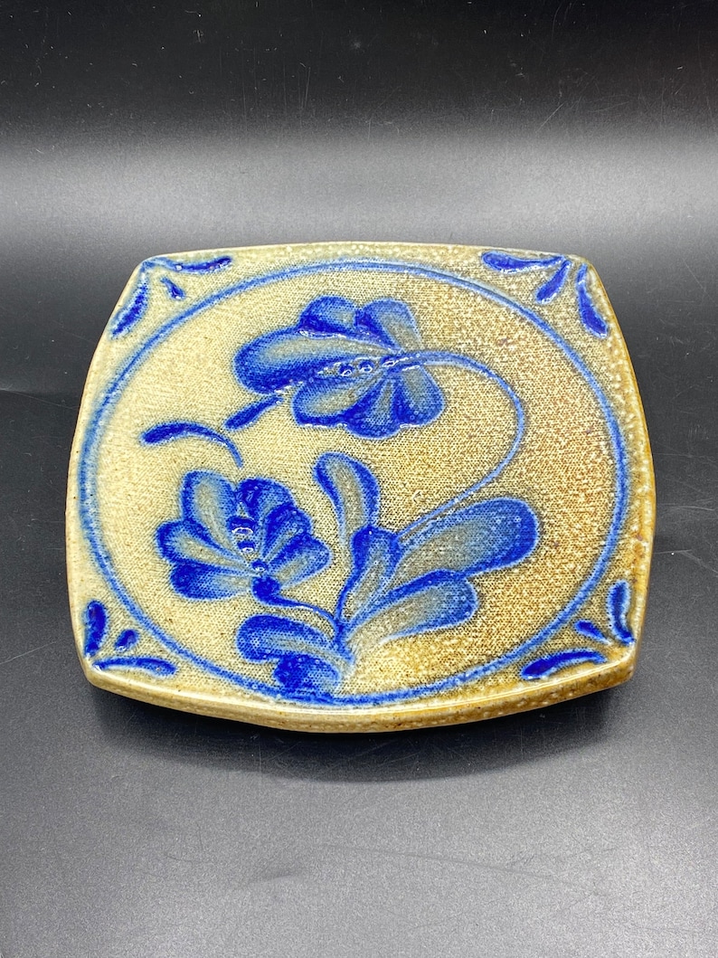 K&ouml;nnte beinhalten: Eine quadratische Keramikfliese mit einem blauen und wei&szlig;en Blumenmuster. Die Fliese hat eine strukturierte Oberfl&auml;che und einen leicht erh&ouml;hten Rand.