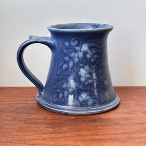 Può includere: Una tazza di ceramica blu con manico e motivo floreale in rilievo. La tazza ha una base ampia che si assottiglia verso l'alto, con un bordo svasato. Il manico è curvo e attaccato al lato. La tazza è su una superficie di legno.