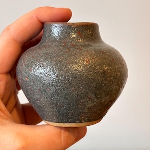 May include: A small, speckled ceramic vase with a rounded body and a narrow neck. The vase has a dark gray base color with reddish-brown speckles. The rim and base are a lighter, natural clay color. The vase is held in a hand.