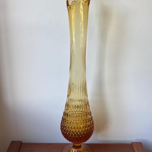 May include: A tall, amber-coloured glass vase featuring a flared, scalloped rim and a long, slender neck with vertical ribbing. The base of the vase has a bulbous shape with a textured, hobnail-like pattern, supported by a circular pedestal foot.