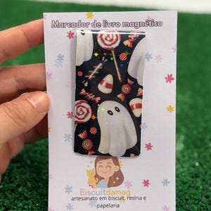 Pode incluir: Marcador de livros magnético retangular com design de Halloween. O design apresenta fantasmas brancos, doces de milho e pirulitos em um fundo preto. O marcador está preso a um cartão branco com texto em português.