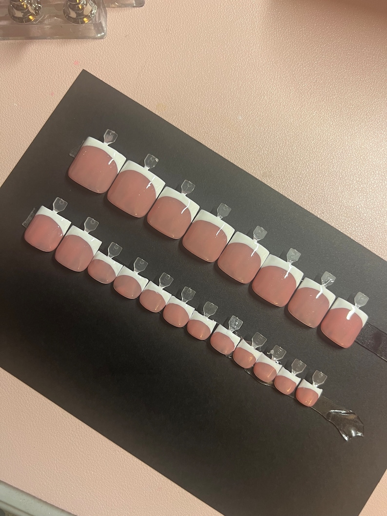 May include: A set of 14 false nails with a white tip and a pink base. The nails are arranged in two rows on a black surface.