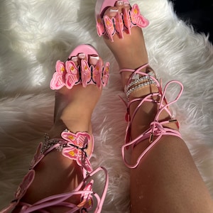 May include: Pink high-heeled sandals with butterfly embellishments and lace-up closures. The sandals have a platform sole and a delicate butterfly design on the straps.