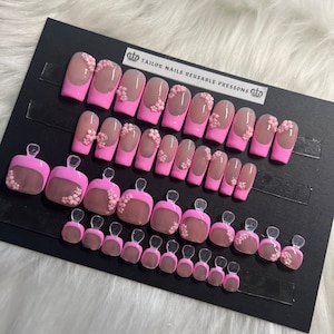 Op de afbeelding: Een set van 24 herbruikbare press-on nagels in roze met een bloemenpatroon. De nagels zijn gerangschikt in rijen op een zwarte displaykaart met de tekst "TAILOR NAILS REUSABLE PRESSONS".
