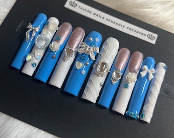 May include: A set of long, reusable press-on nails in a variety of designs. The nails feature a light blue base, white accents, and glittery pink tips. Embellishments include bows, hearts, and floral designs with rhinestones.