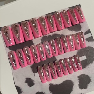 May include: A set of pink press-on nails with a rectangular shape. Each nail features a pink base, a clear center, and is adorned with silver rhinestones. The nails are arranged on a patterned surface.
