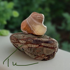 Pode incluir: Uma escultura em pedra polida. A base &eacute; uma pedra escura e oblonga com estrias castanhas e cinzentas. Uma pedra menor e mais clara com faixas laranja e brancas est&aacute; colocada em cima. O fundo &eacute; verde desfocado.