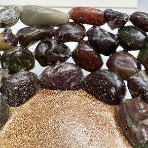 May include: An assortment of polished stones in various colors, including brown, green, and speckled patterns. The stones are arranged on a bed of small, tan-colored granules, creating a natural, textured display. The stones vary in size and shape.