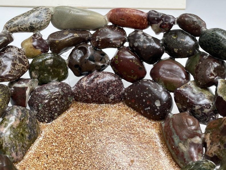 May include: An assortment of polished stones in various colors, including brown, green, and speckled patterns. The stones are arranged on a bed of small, tan-colored granules, creating a natural, textured display. The stones vary in size and shape.