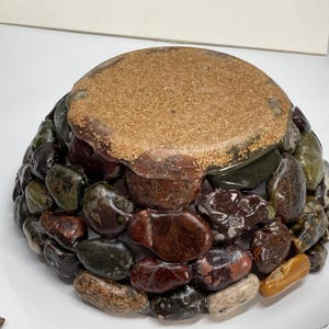 May include: A decorative bowl with a rounded shape, crafted from a mosaic of polished stones in shades of brown, green, and red. The top surface is a light brown, textured material. The bowl is designed to hold small items.