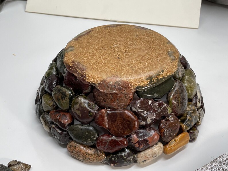 May include: A decorative bowl with a rounded shape, crafted from a mosaic of polished stones in shades of brown, green, and red. The top surface is a light brown, textured material. The bowl is designed to hold small items.
