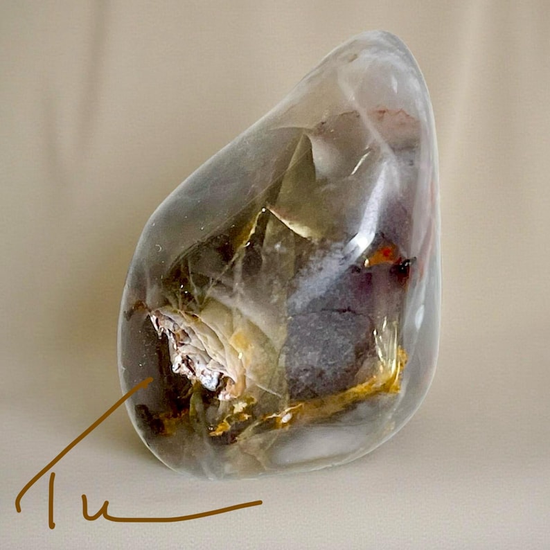 Pode incluir: Uma pedra polida e transl&uacute;cida com formato arredondado. A pedra apresenta padr&otilde;es internos brancos, castanhos e dourados, com toques de vermelho e roxo. A palavra "Tu" est&aacute; escrita em it&aacute;lico no canto inferior esquerdo.