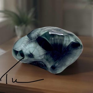 Pode incluir: Um objeto decorativo transl&uacute;cido de forma irregular, com uma mistura de cores verde-azulado escuro e claro. O objeto tem uma superf&iacute;cie lisa e polida e parece ser feito de vidro ou material semelhante. A palavra "Tu" est&aacute; escrita em it&aacute;lico no canto inferior esquerdo.