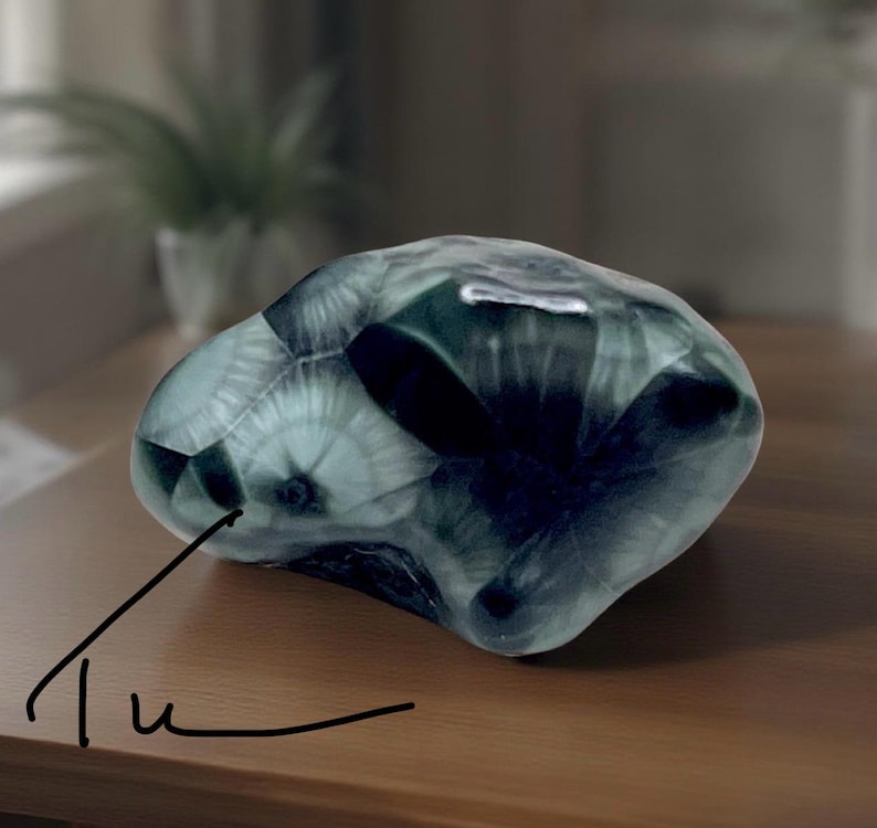 Pode incluir: Um objeto decorativo transl&uacute;cido de forma irregular, com uma mistura de cores verde-azulado escuro e claro. O objeto tem uma superf&iacute;cie lisa e polida e parece ser feito de vidro ou material semelhante. A palavra "Tu" est&aacute; escrita em it&aacute;lico no canto inferior esquerdo.