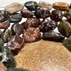 May include: A collection of polished stones in various colors, including brown, green, and red, arranged around a bed of small, tan grains. The stones have a smooth, rounded shape and a natural, earthy appearance. The arrangement creates a visually appealing display.