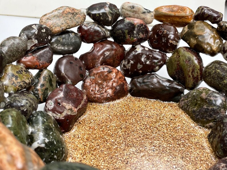 May include: A collection of polished stones in various colors, including brown, green, and red, arranged around a bed of small, tan grains. The stones have a smooth, rounded shape and a natural, earthy appearance. The arrangement creates a visually appealing display.