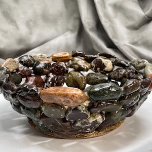 May include: A decorative bowl crafted from polished stones in shades of brown, green, and tan. The bowl's exterior is constructed with a mosaic of stones, creating a textured surface. The interior is filled with similar stones, offering a natural, earthy aesthetic.