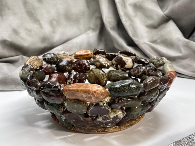 May include: A decorative bowl crafted from polished stones in shades of brown, green, and tan. The bowl's exterior is constructed with a mosaic of stones, creating a textured surface. The interior is filled with similar stones, offering a natural, earthy aesthetic.
